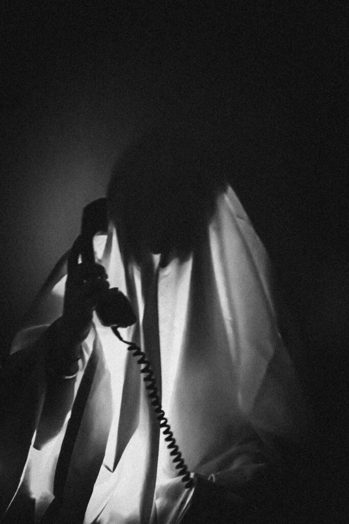 A mysterious figure shrouded in a sheet holds a vintage telephone in a dark setting.
